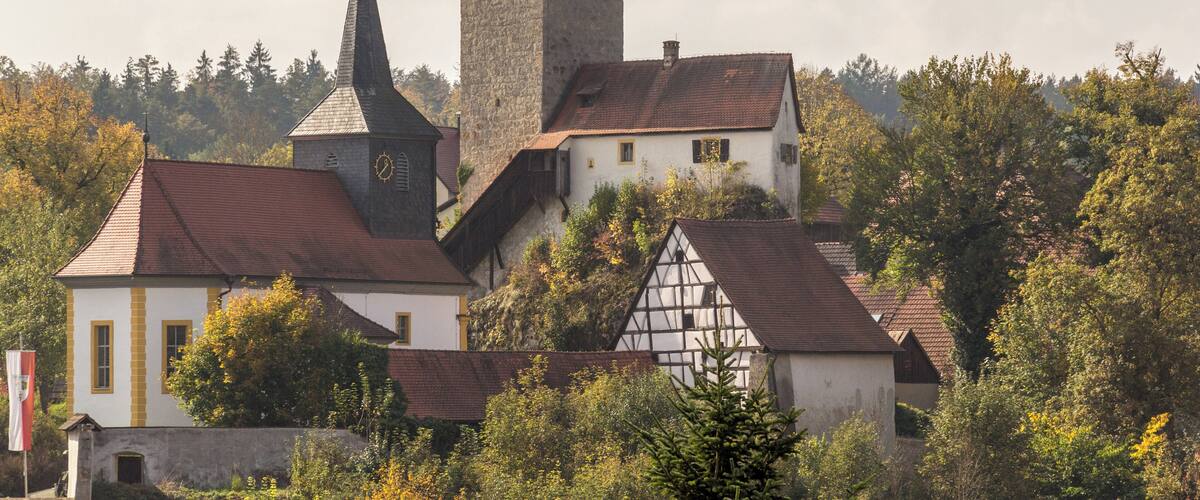 A church with a tall tower and a smaller steeple, surrounded by trees and traditional buildings.