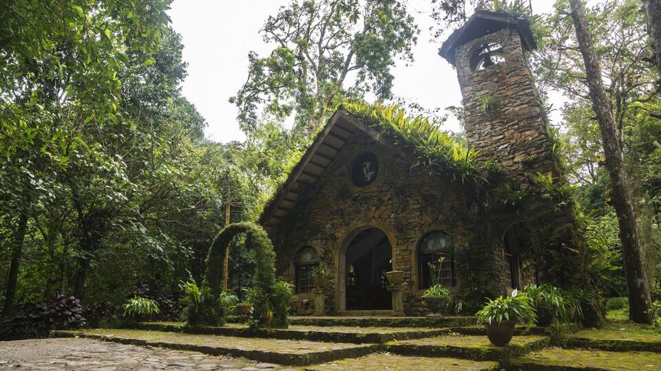 A stone building with a tower, surrounded by lush greenery and a cobblestone path.