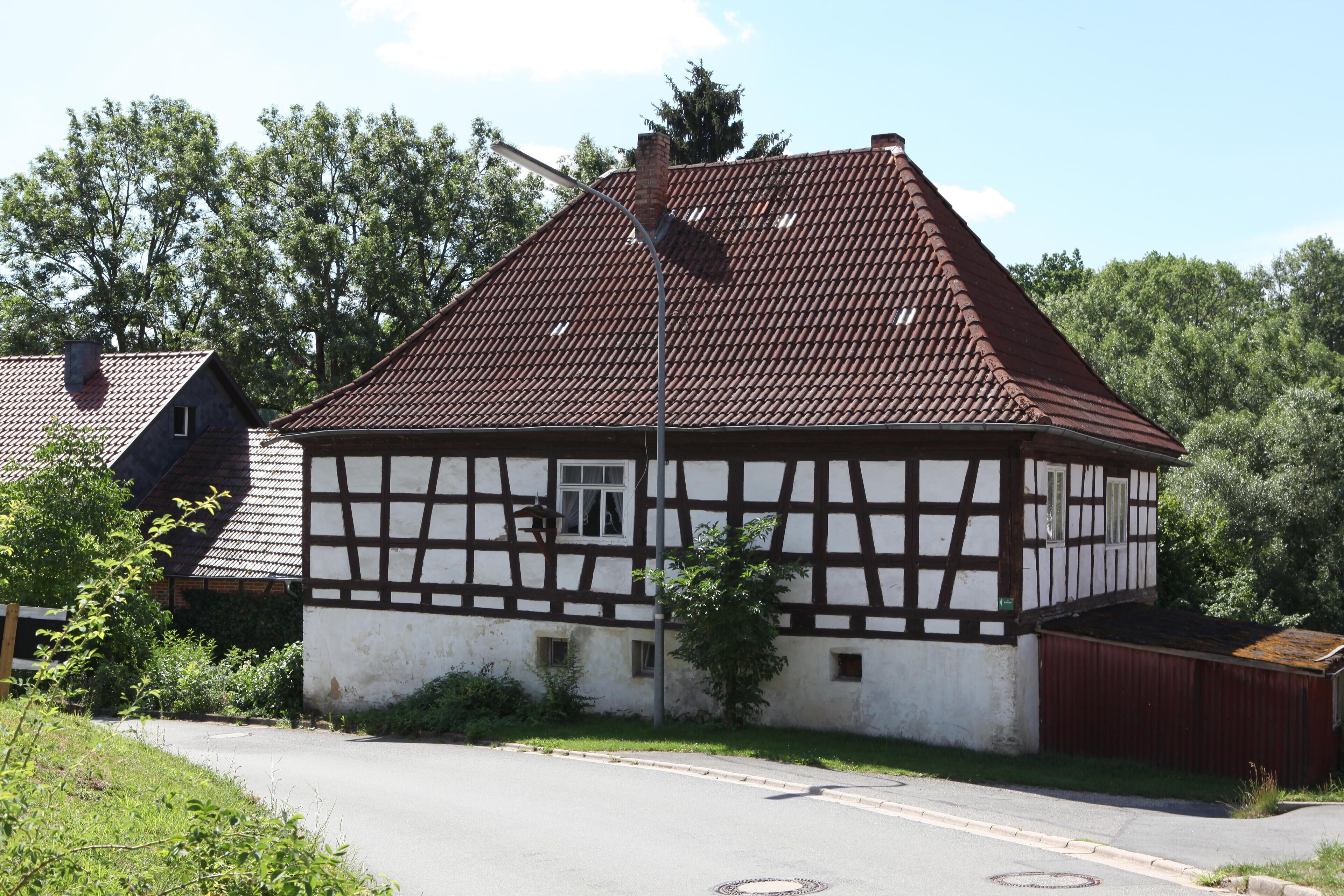 A traditional half-timbered house with a sloped roof, surrounded by greenery and a clear sky.