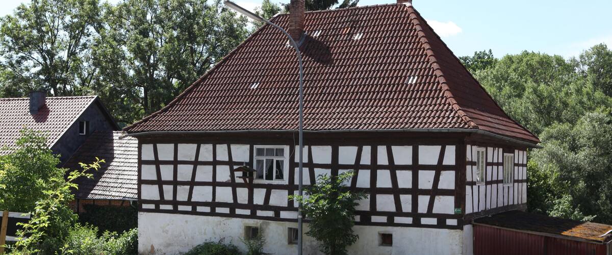 A traditional half-timbered house with a sloped roof, surrounded by greenery and a clear sky.