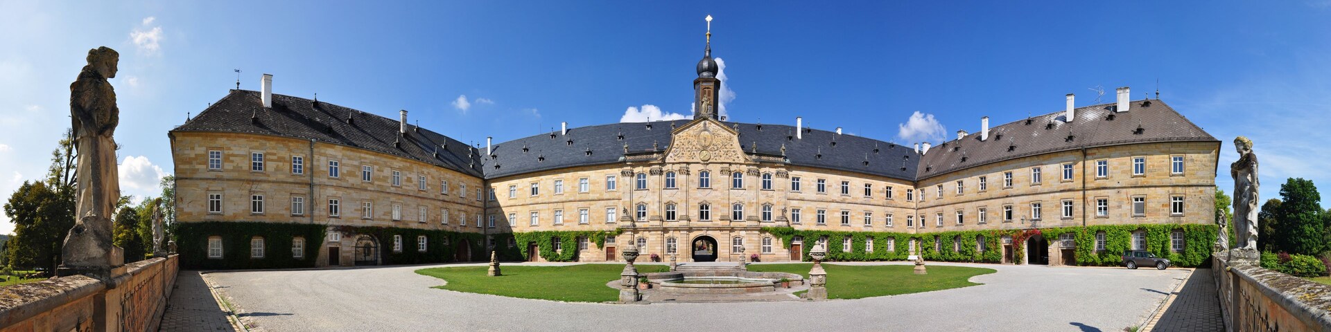 Un grand bâtiment historique doté d’une tour centrale et de deux ailes symétriques, entouré d’un jardin bien entretenu et d’une cour dallée.