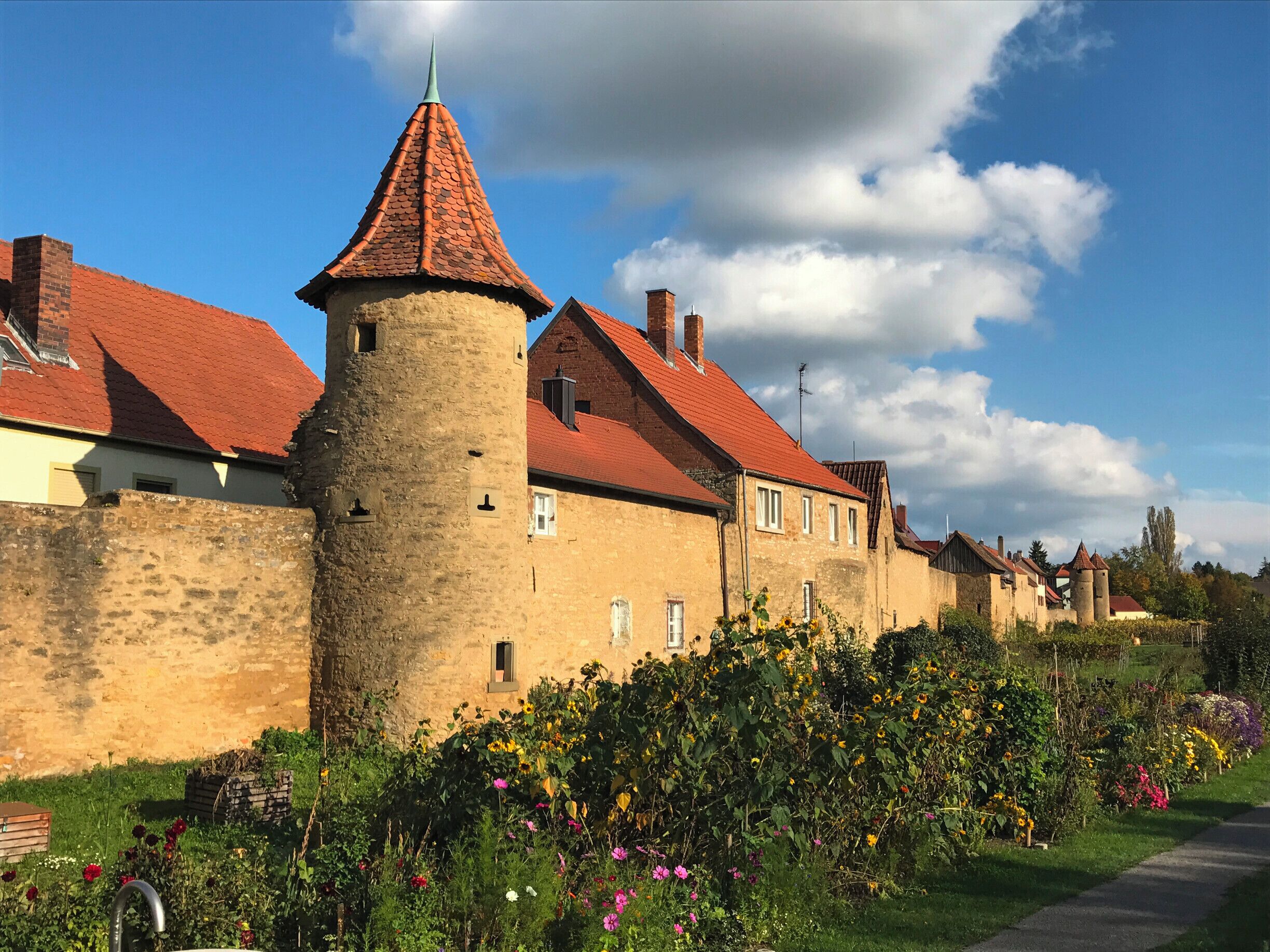 A historic stone wall with a tower, red-roofed buildings, and a garden with colorful flowers.