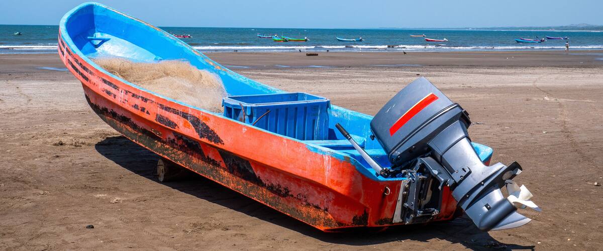 A large, rusted boat with a blue interior and a motor, lying on a sandy beach with the ocean in the background.