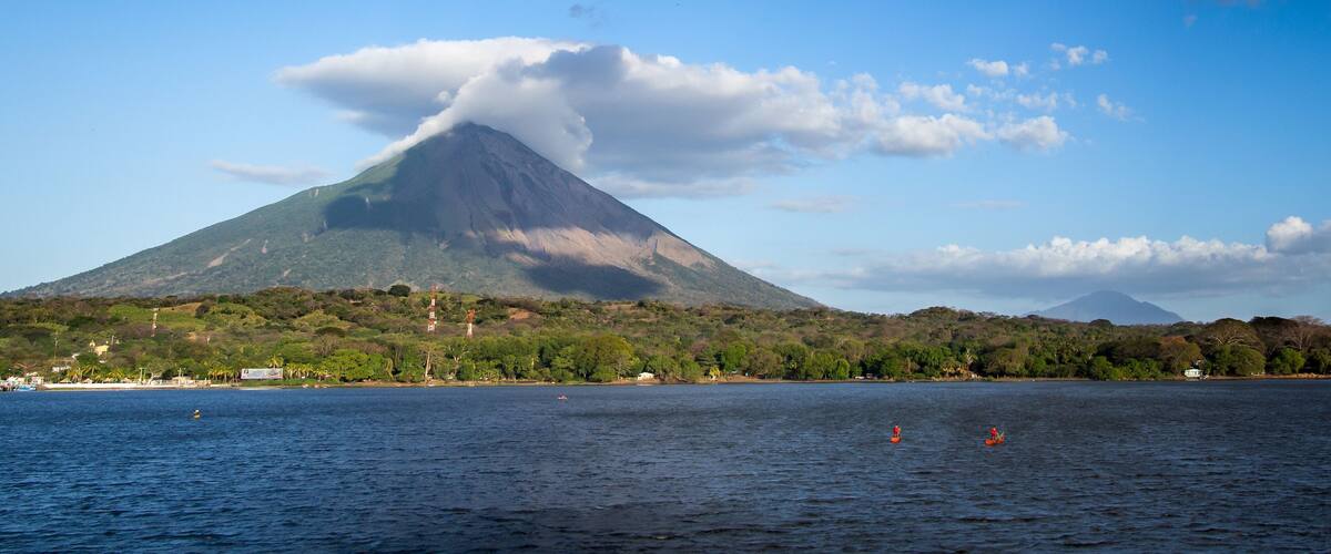 A volcano with a cloud of smoke on its peak, a body of water with boats, and a shoreline with buildings and trees.