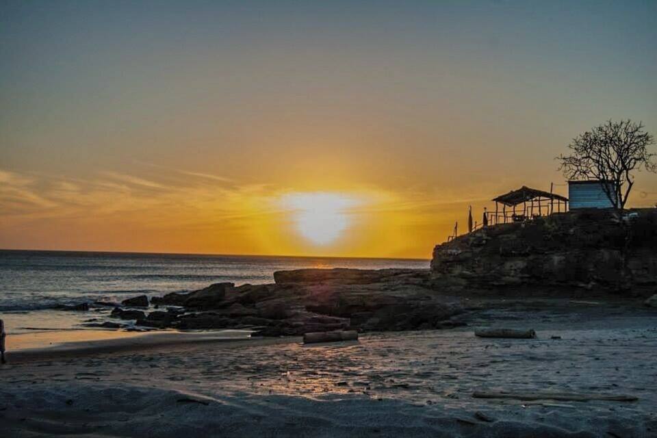 Un coucher de soleil sur une plage rocheuse, avec une petite construction sur la droite.