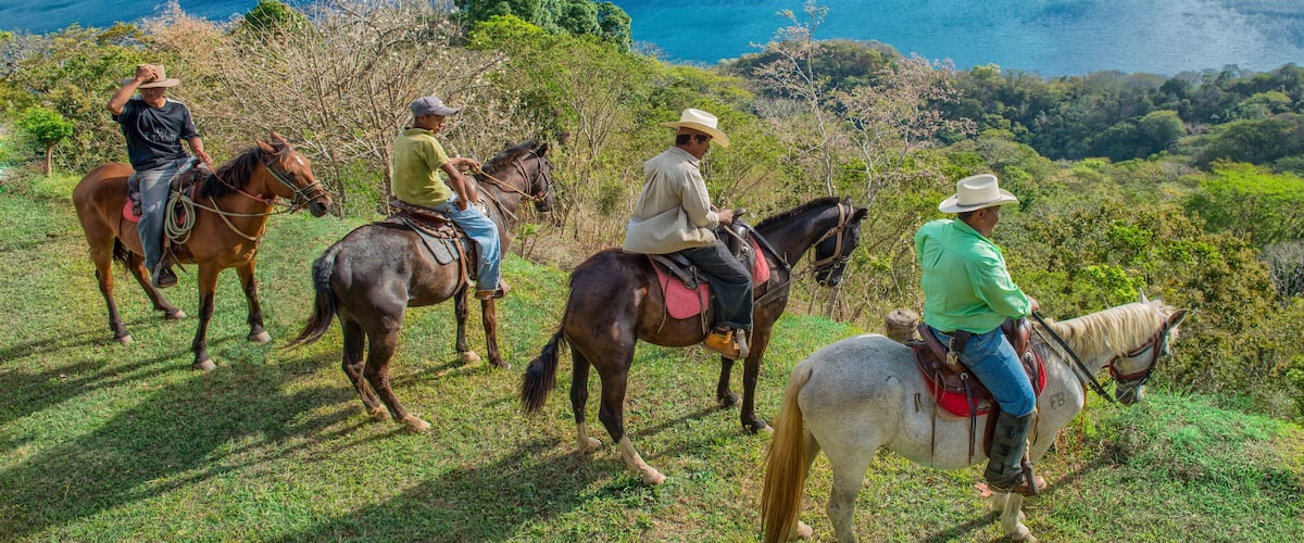 A group of people riding horses on a grassy hillside with a view of a large body of water and cliffs in the background.