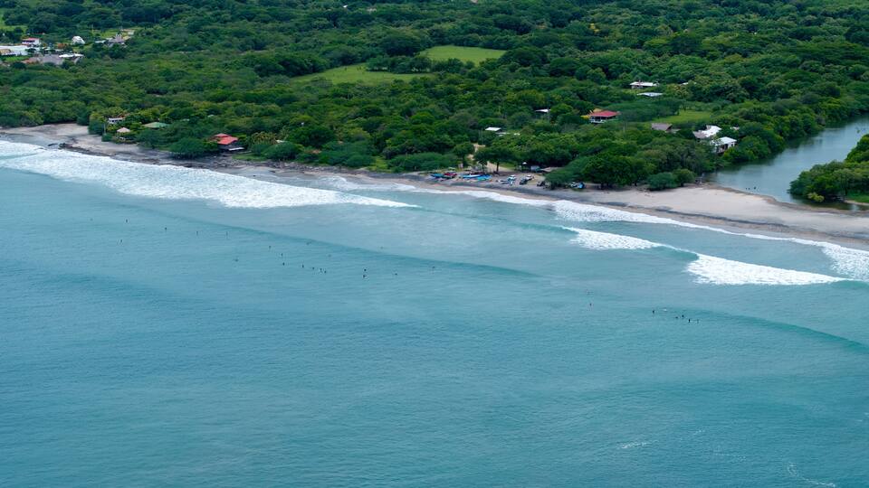 A coastal area with a beach, ocean waves, and a forested hillside with scattered buildings.