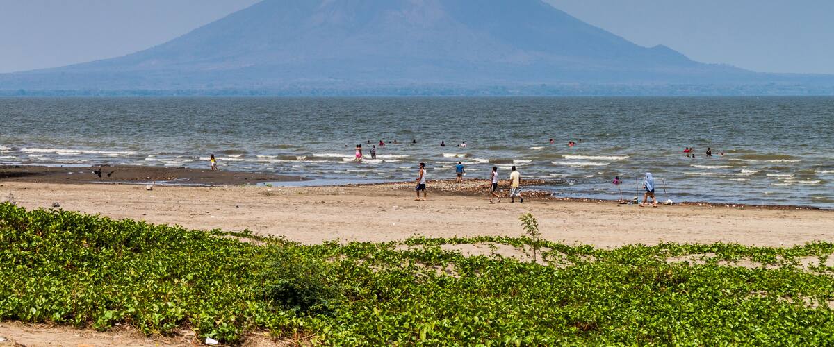 A beach with people swimming and walking, a volcano in the background, and green vegetation in the foreground.