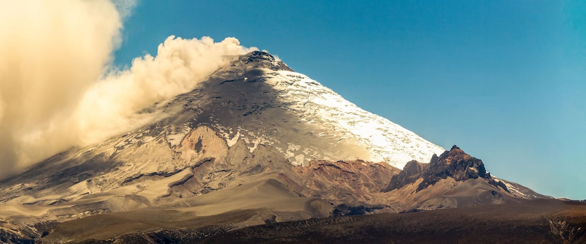 A snow-capped mountain with a prominent peak and a cloud of dust or smoke rising from its base.