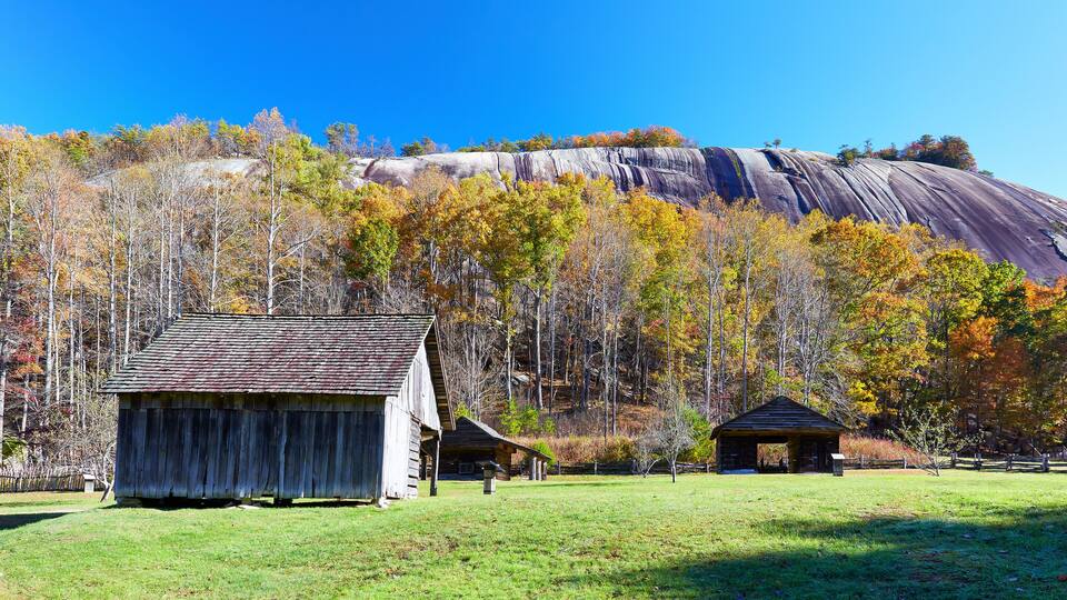 A rustic wooden cabin in a grassy field with a forest and a large rock formation in the background.