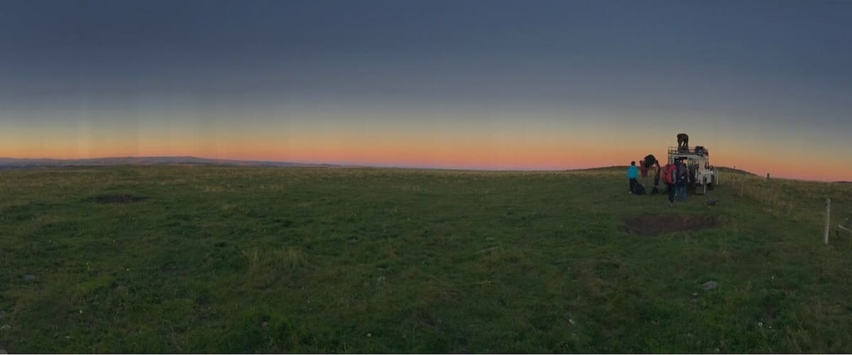 A grassy field with a sunset in the background and a group of people near a vehicle.