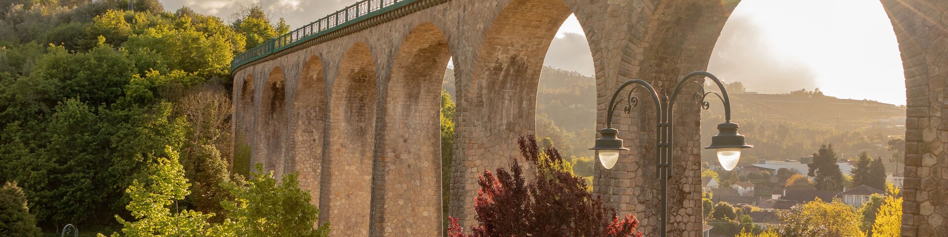 Uma ponte de pedra com grades verdes, arcos e uma paisagem ensolarada com árvores e um parque.