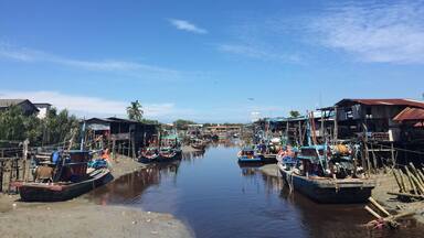 A calm river with boats docked on both sides, surrounded by small buildings and palm trees.