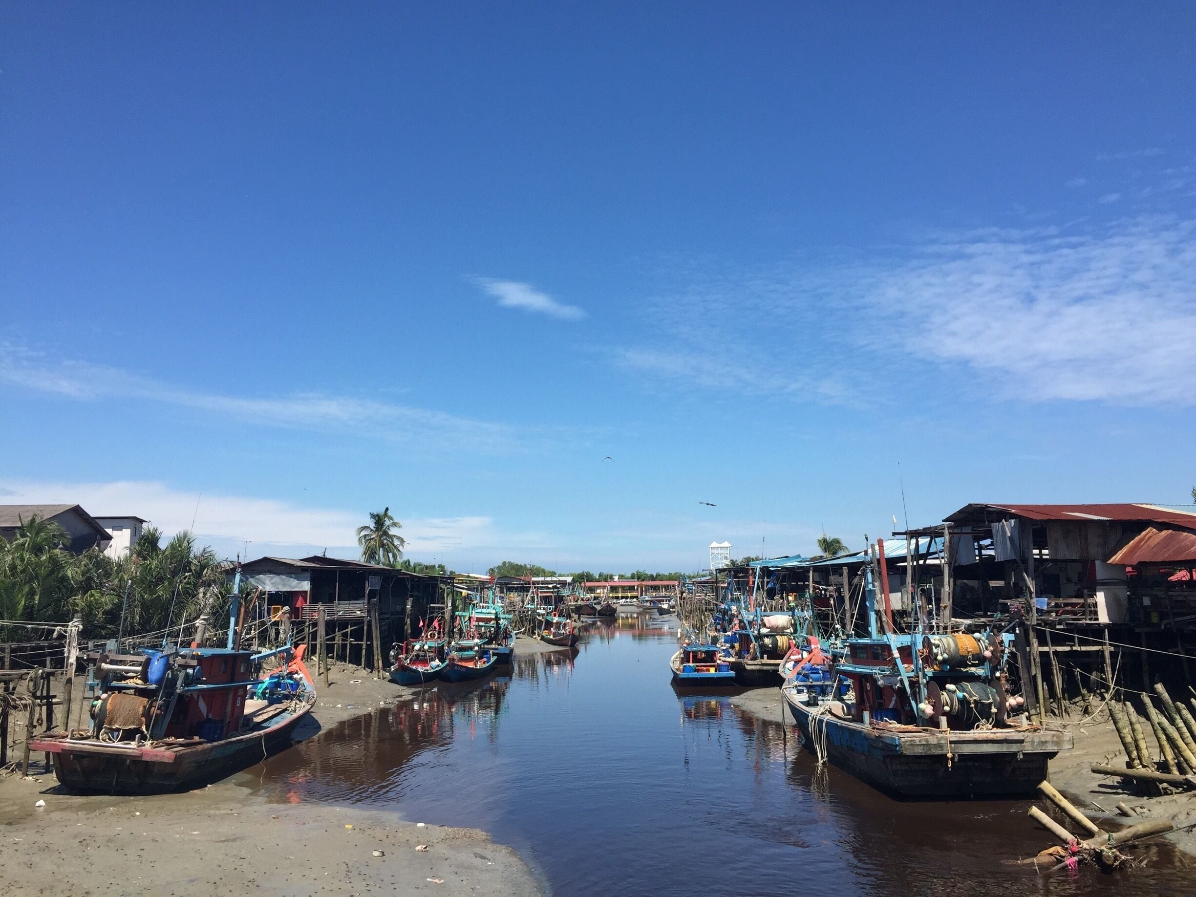A calm river with boats docked on both sides, surrounded by small buildings and palm trees.