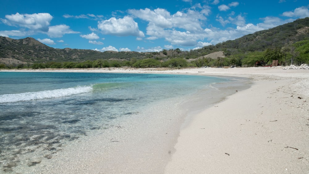 A beach with clear turquoise water, white sand, and green hills in the background.