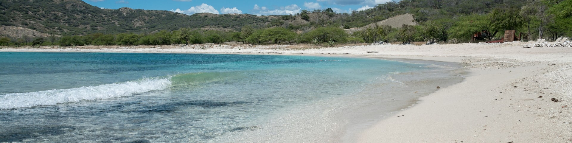 A beach with clear turquoise water, white sand, and green hills in the background.