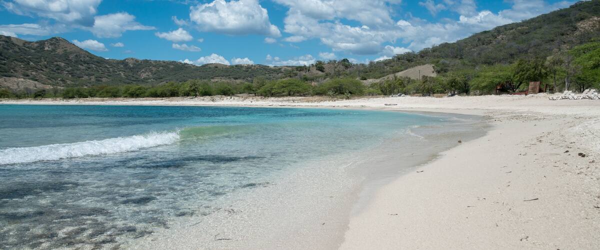 A beach with clear turquoise water, white sand, and green hills in the background.