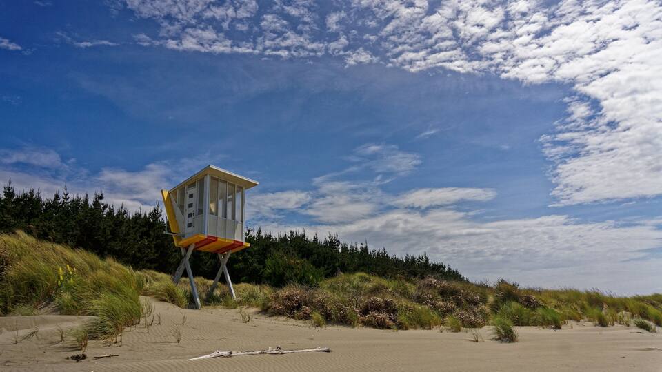 A lifeguard tower on a sandy beach with dunes and coniferous trees in the background.
