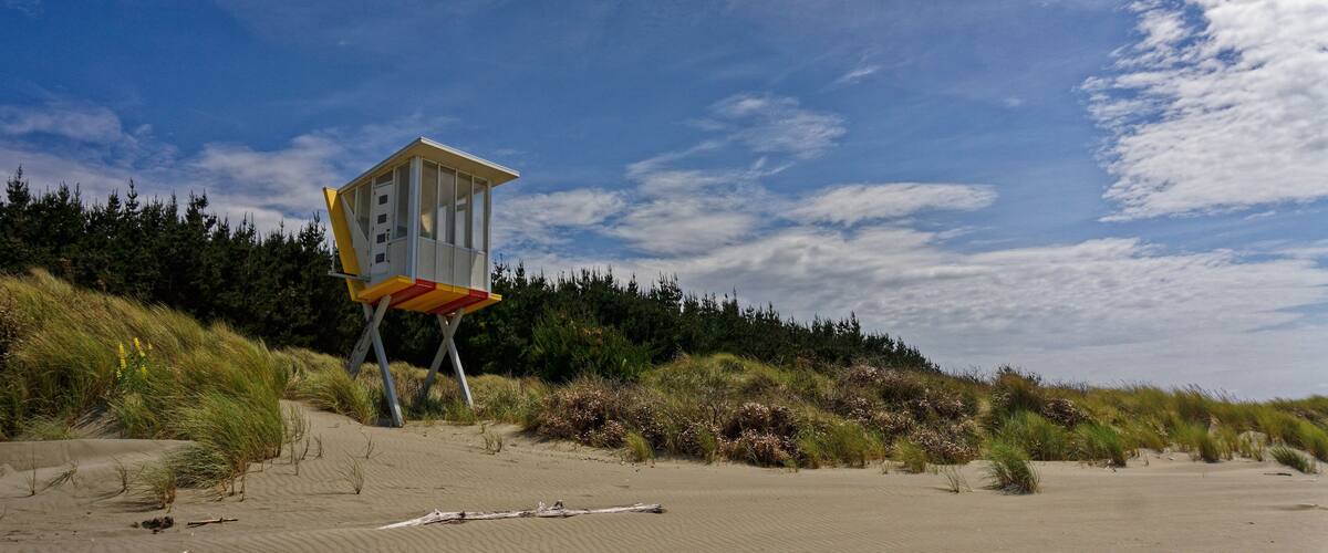 A lifeguard tower on a sandy beach with dunes and coniferous trees in the background.
