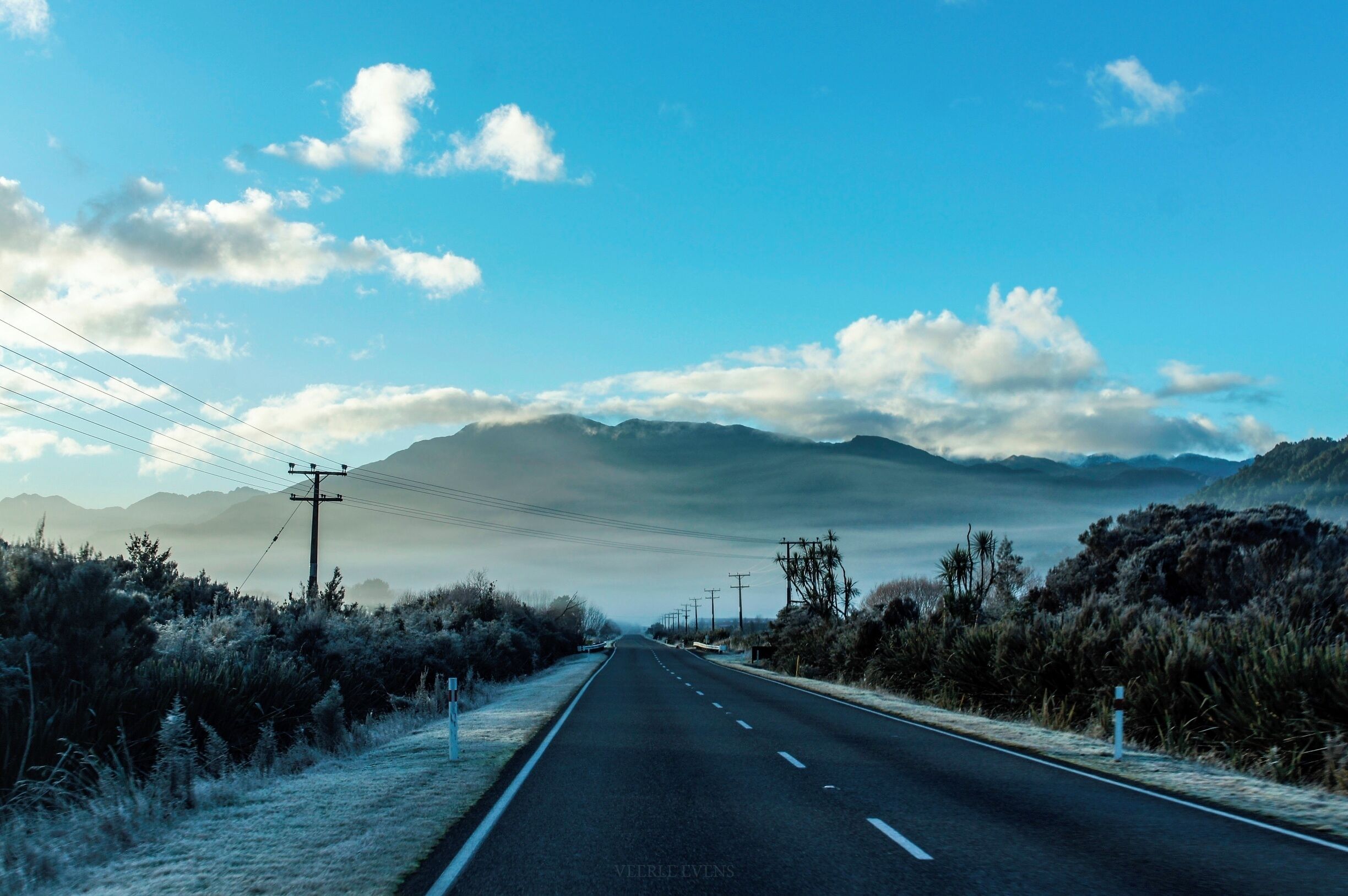 A road flanked by dry bushes and power lines, leading towards distant mountains under a clear sky.