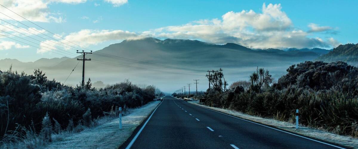 A road flanked by dry bushes and power lines, leading towards distant mountains under a clear sky.