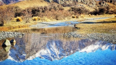 Cordilheira com picos nevados, céu azul límpido e um rio calmo refletindo a paisagem.