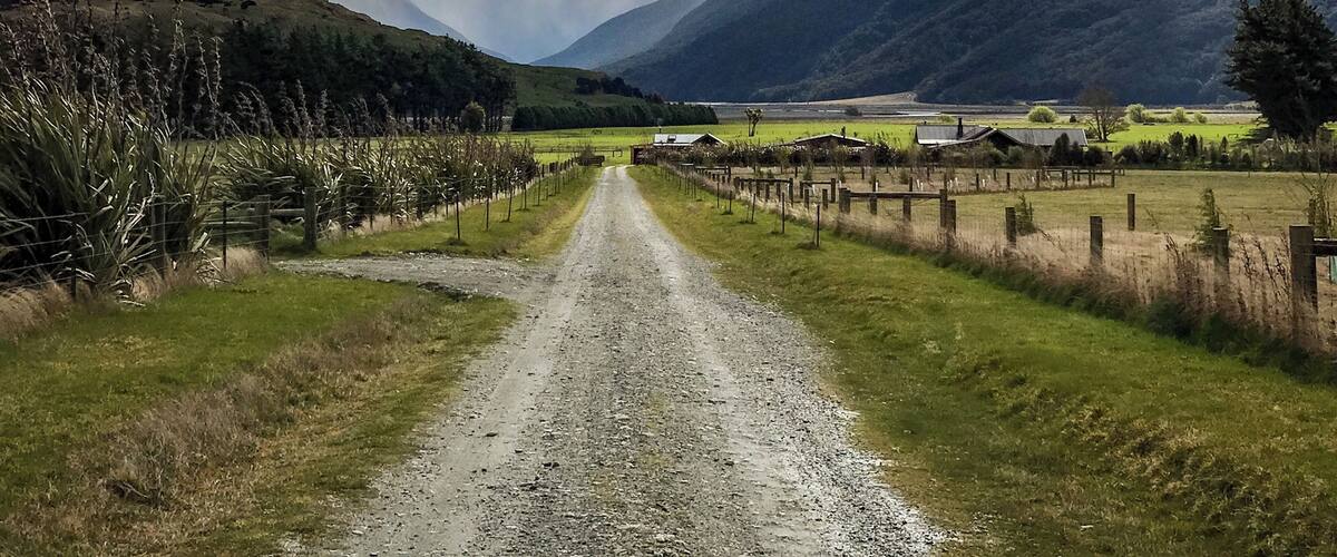 A gravel road leading through a valley with mountains in the background and a cloudy sky.