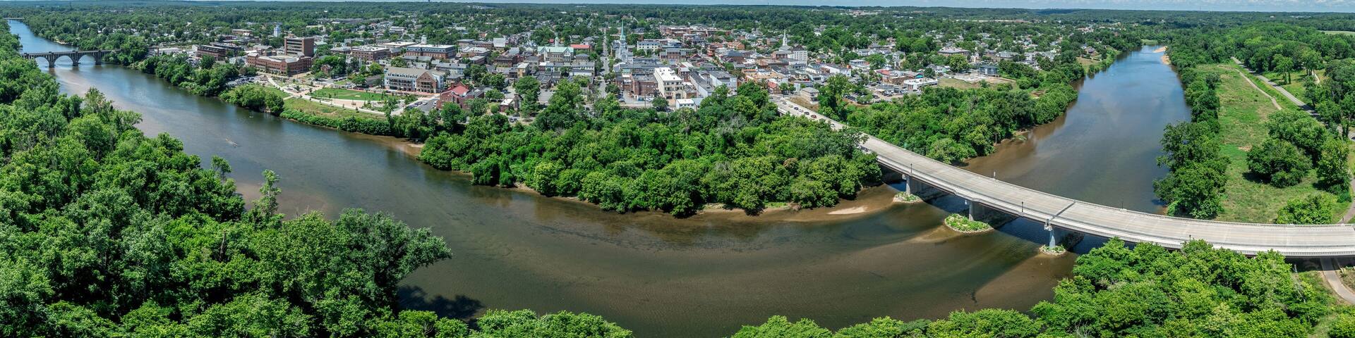 A river flowing through a town with a bridge, surrounded by greenery and a clear blue sky.