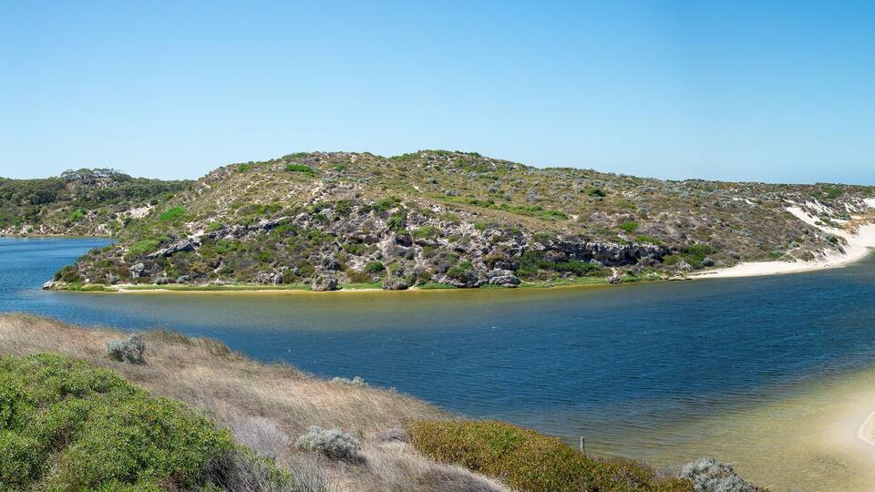 A coastal landscape with a sandy beach, a clear blue sea, and a hill with sparse vegetation.