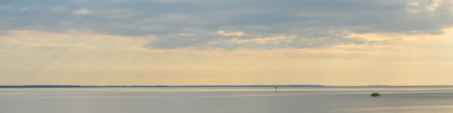 A calm body of water with a distant boat, a grassy foreground, and a cloudy sky.