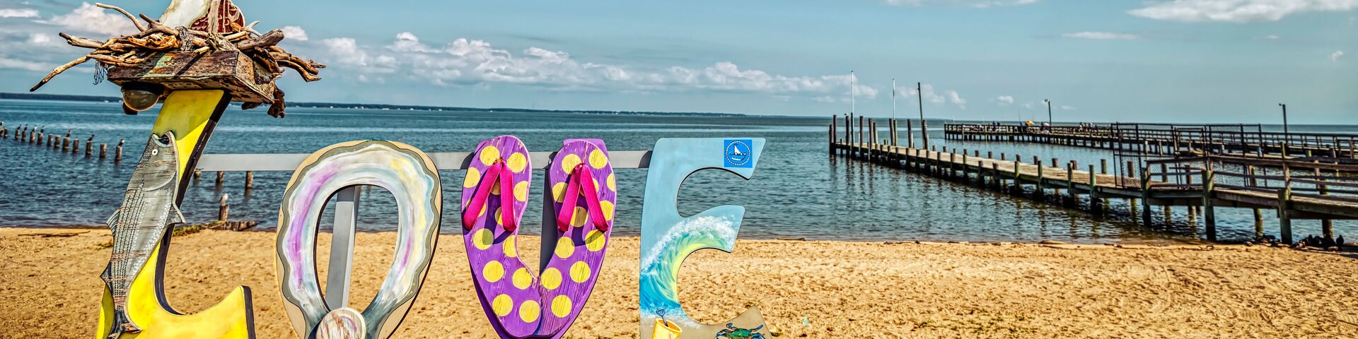 A beach with a wooden bench, colorful flip-flops, and a pier in the background.