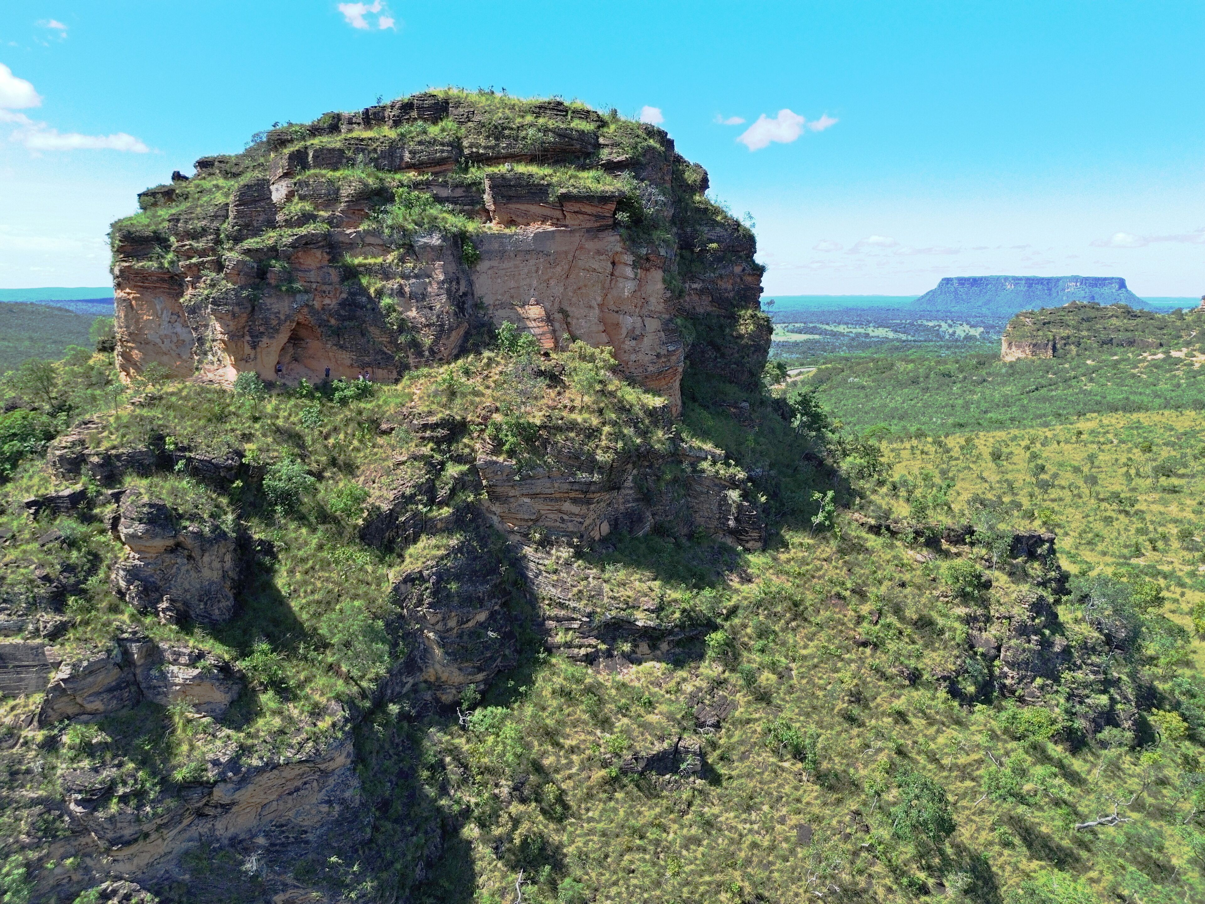 A large rock formation with a flat top, surrounded by green vegetation and a clear blue sky.