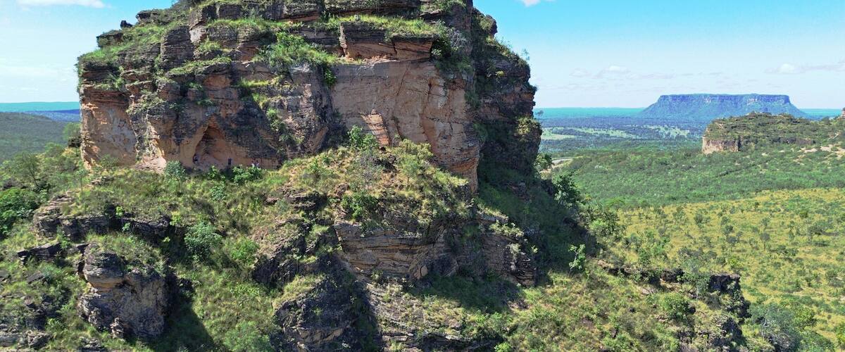 A large rock formation with a flat top, surrounded by green vegetation and a clear blue sky.