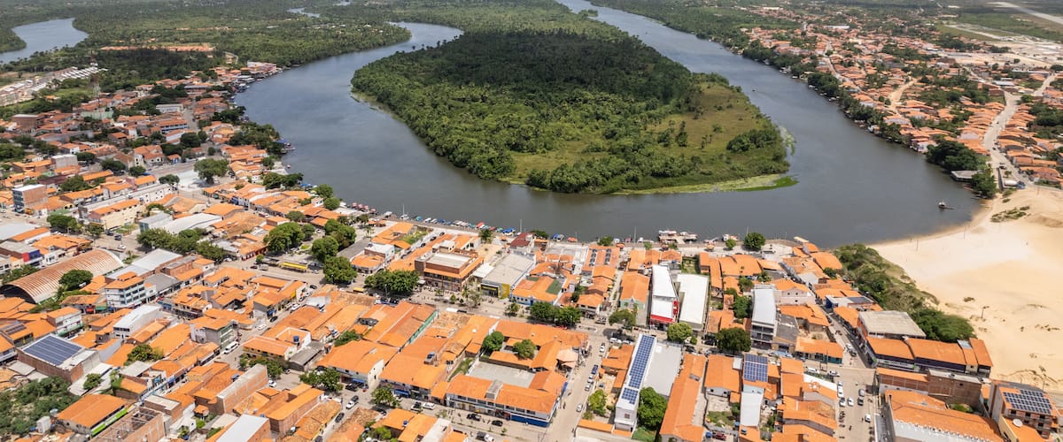An aerial view of a coastal town with a river, green forested island, and orange-roofed buildings.