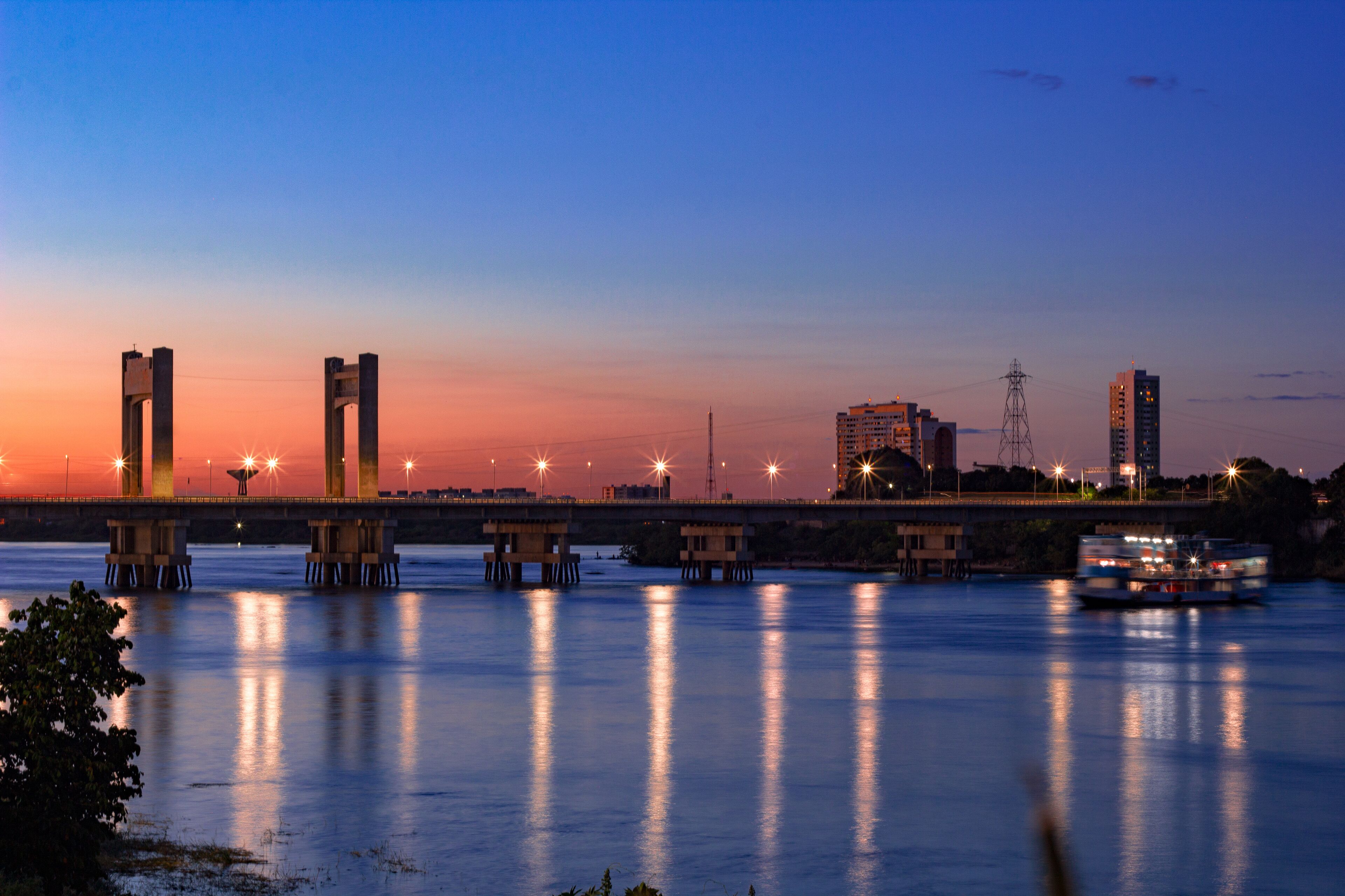 A river at dusk with a bridge, buildings, and a boat.