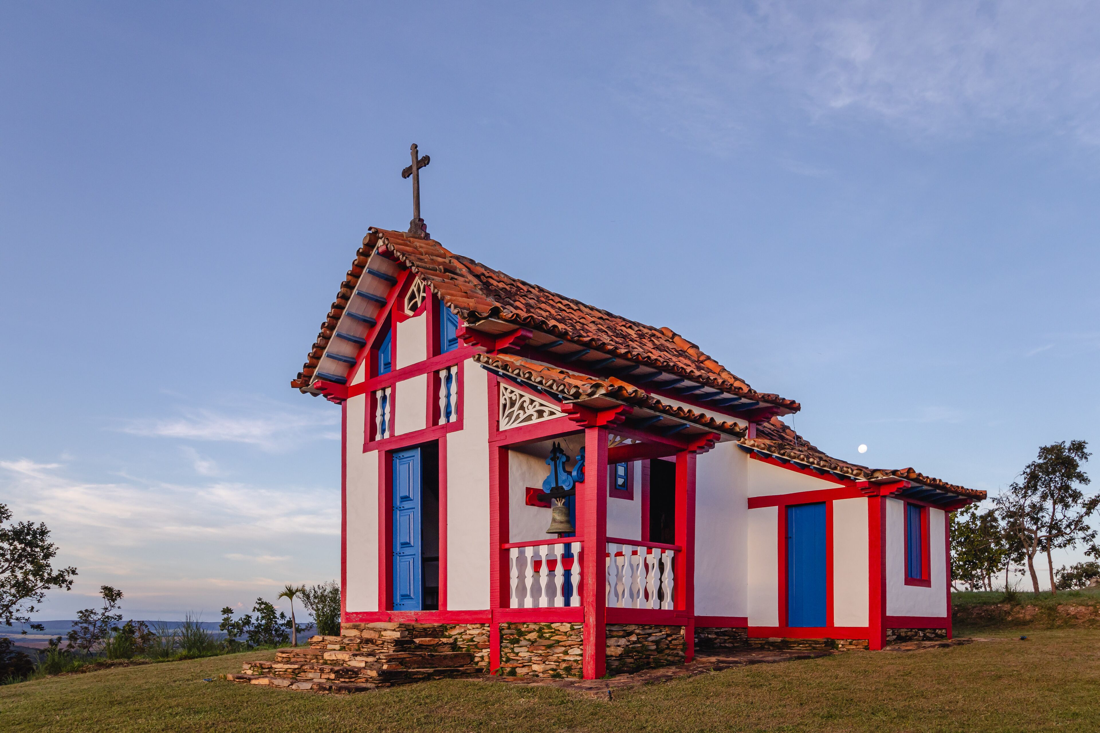 A small church with a red and white facade, a cross on top, and a bell hanging from the entrance.