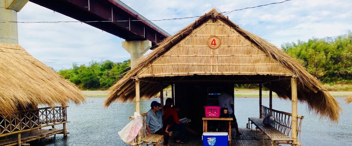 A thatched-roof hut on stilts over water, with a bridge in the background.