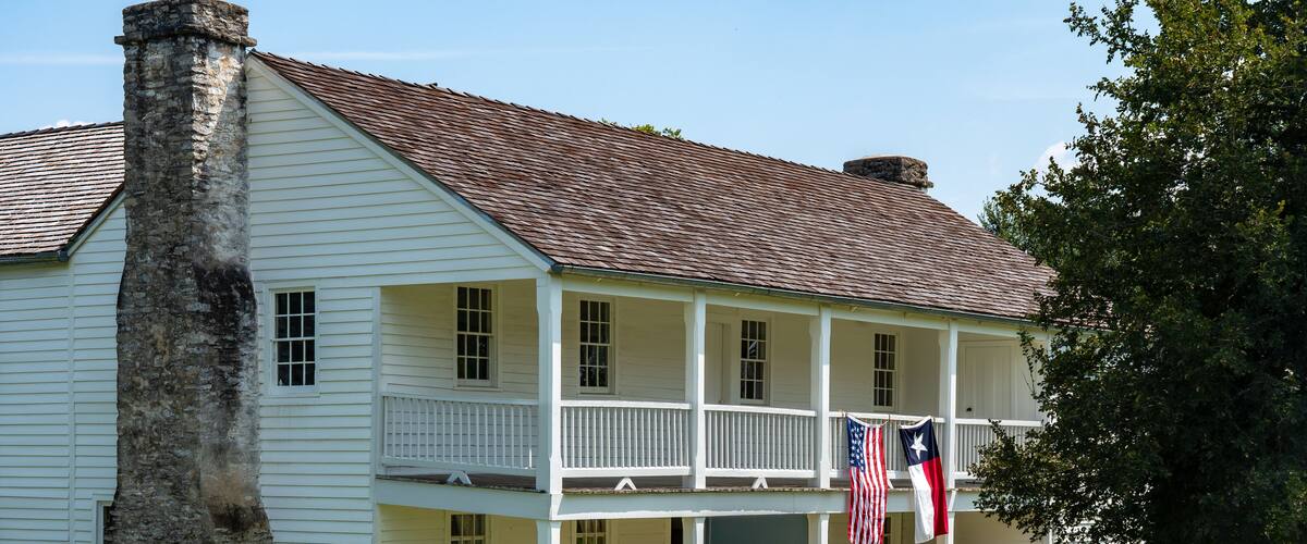 A white colonial-style house with a chimney, a porch, and two flags.