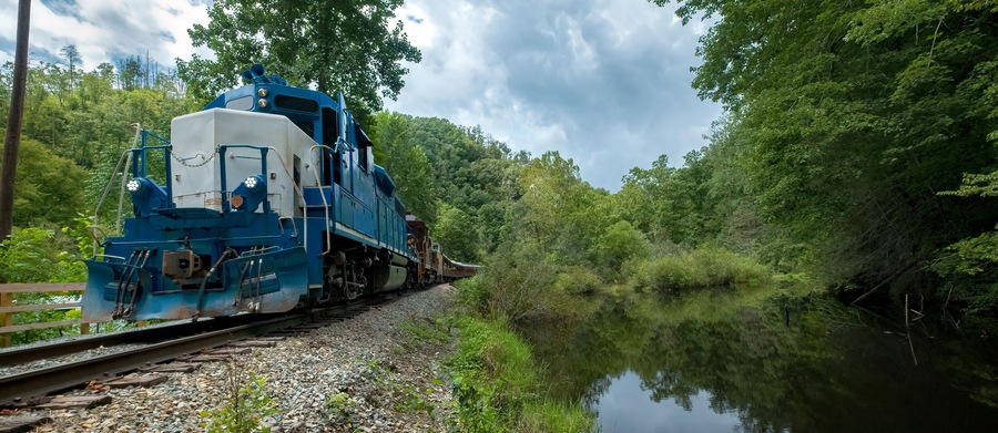 Um trem azul em uma linha estreita, cercado por uma floresta densa e um rio calmo.