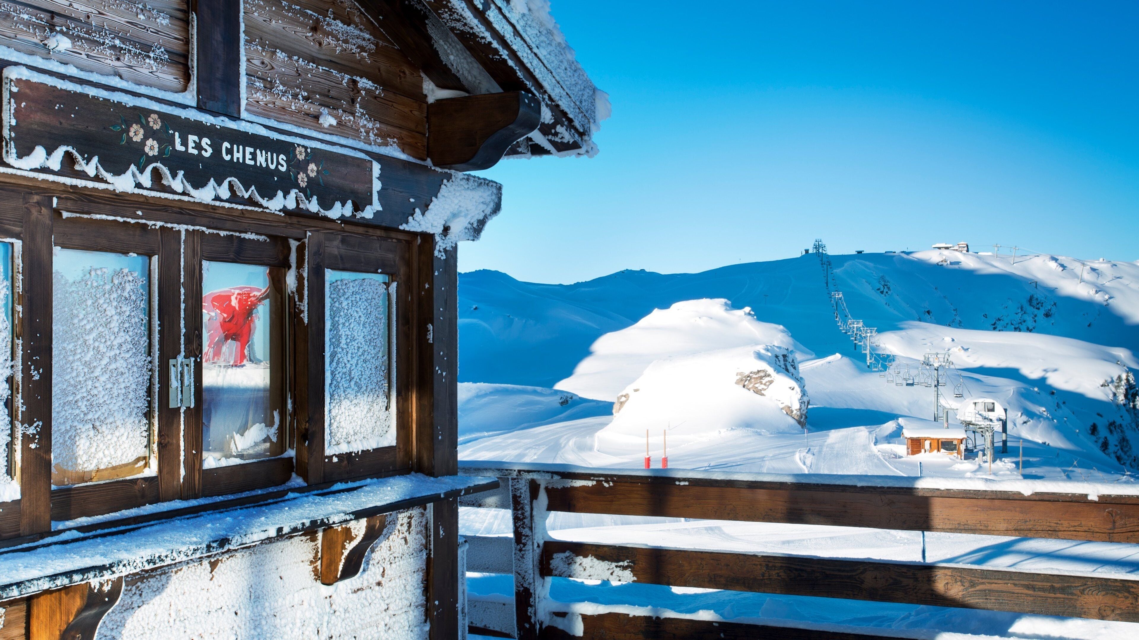A wooden cabin with a sign reading "LES CHENUS" and a snowy mountain landscape in the background.