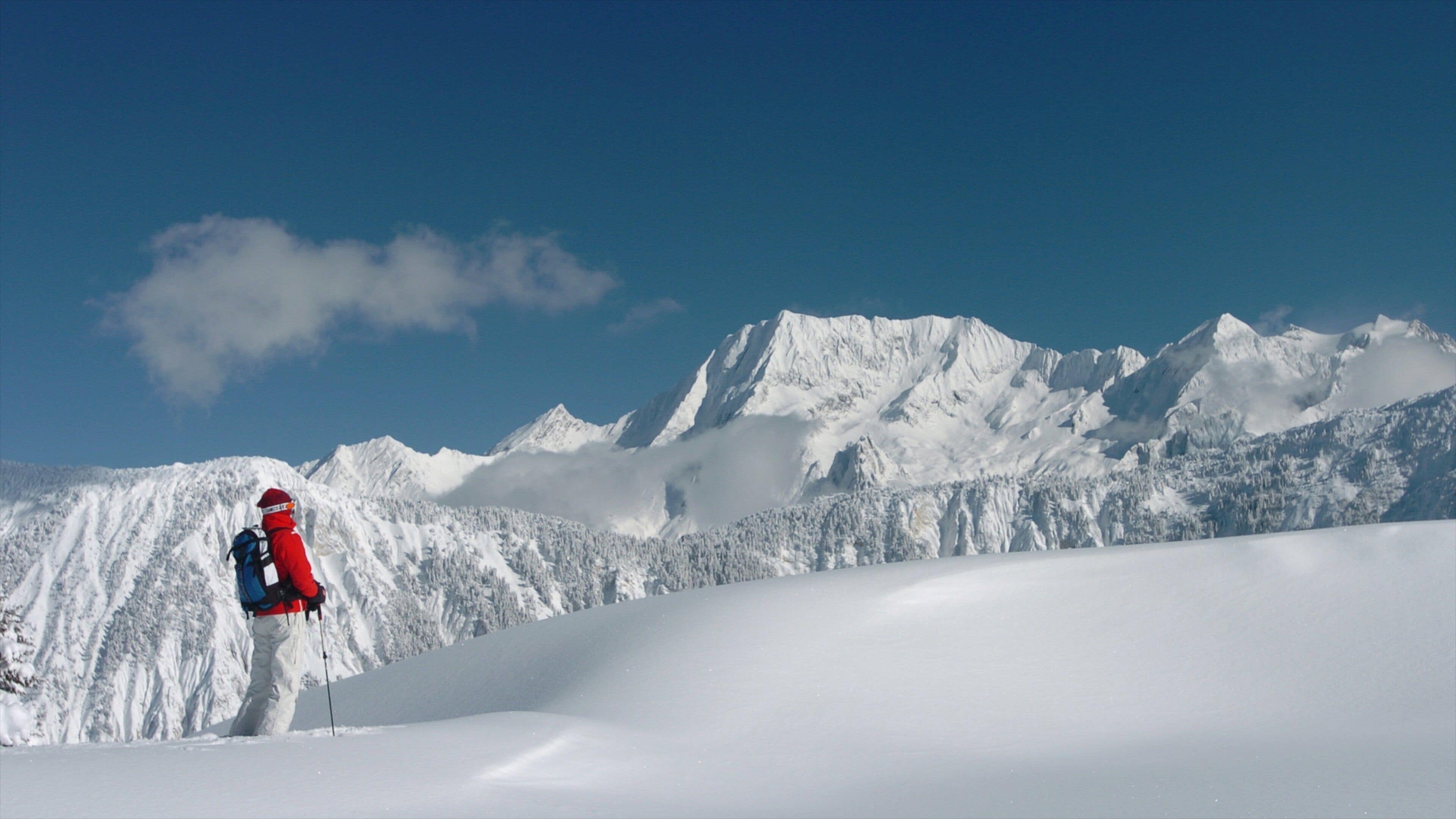 Una persona con equipo rojo y blanco de pie en una pendiente nevada, con una cadena montañosa al fondo.