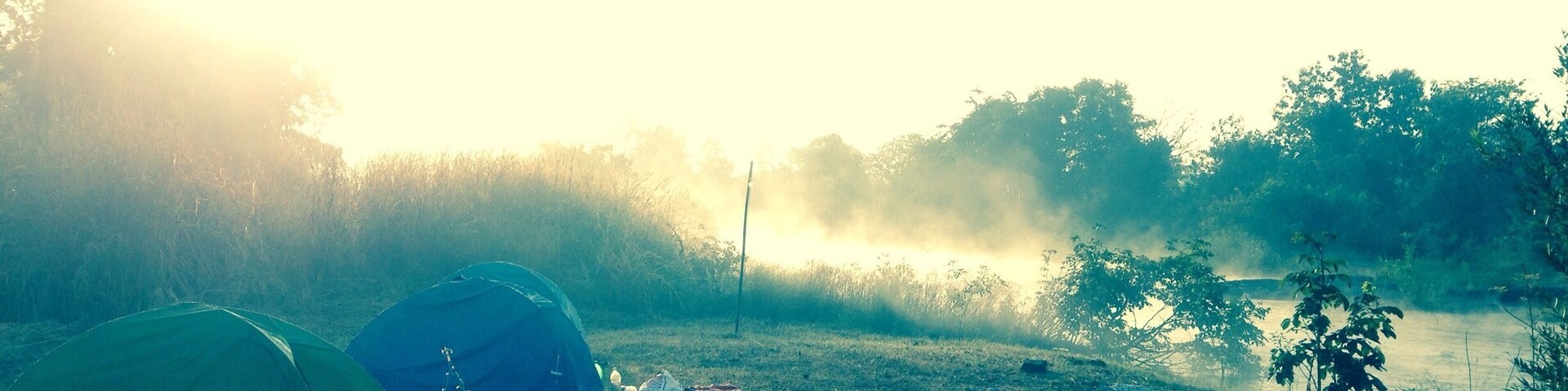 Dos tiendas de campaña montadas en un campo cubierto de hierba cerca de un río, con árboles y niebla al fondo.