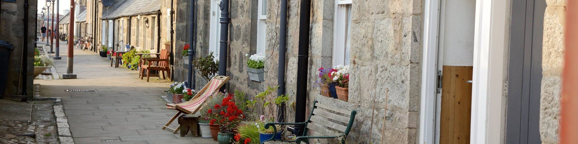 A cobblestone street lined with stone buildings, outdoor seating, and potted plants.