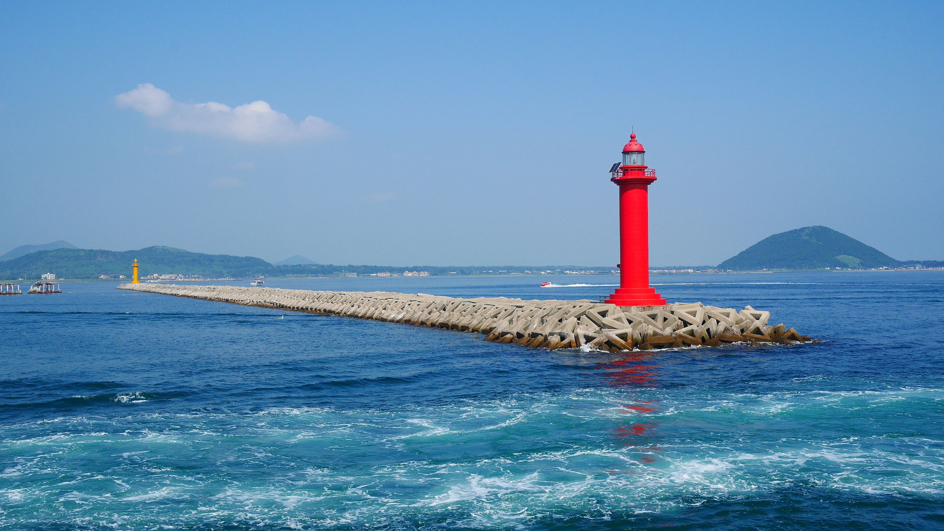 A red lighthouse on a pier extending into the sea, with calm waters and a clear sky.