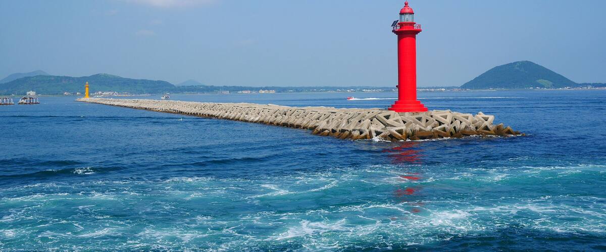 A red lighthouse on a pier extending into the sea, with calm waters and a clear sky.