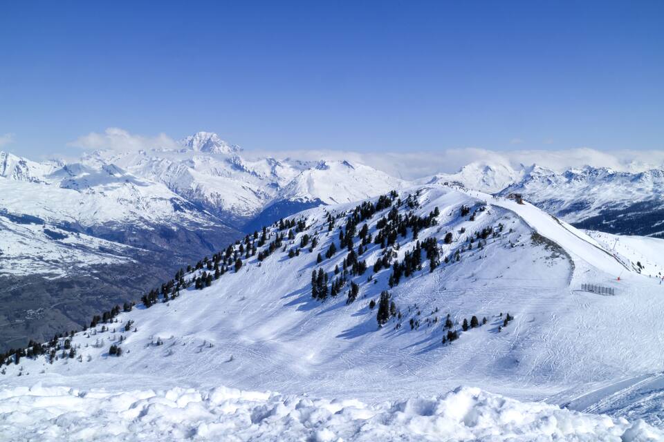A snow-covered mountain slope with a ski trail, trees, and distant peaks under a clear blue sky.