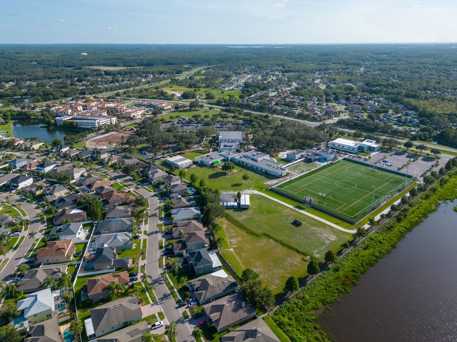 An aerial view of a residential area with houses, a large sports field, and a body of water.