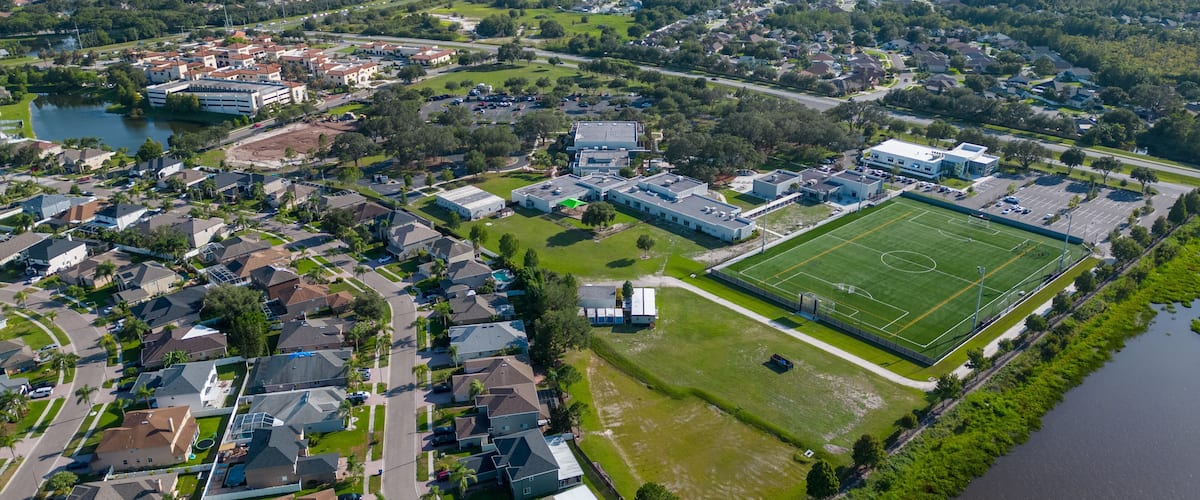 An aerial view of a residential area with houses, a large sports field, and a body of water.
