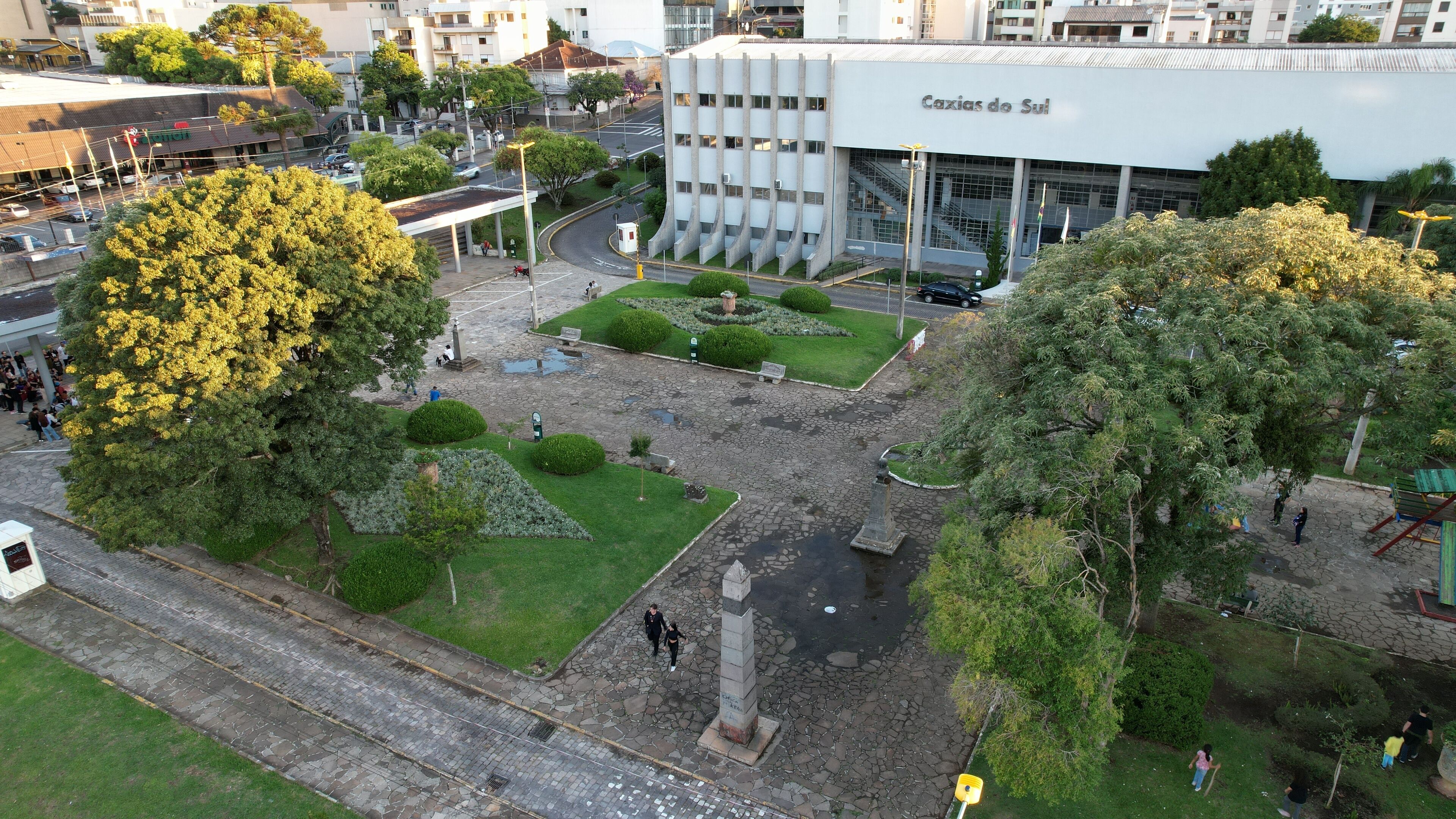 A plaza with a monument, surrounded by trees and a building labeled "Caxias do Sul."