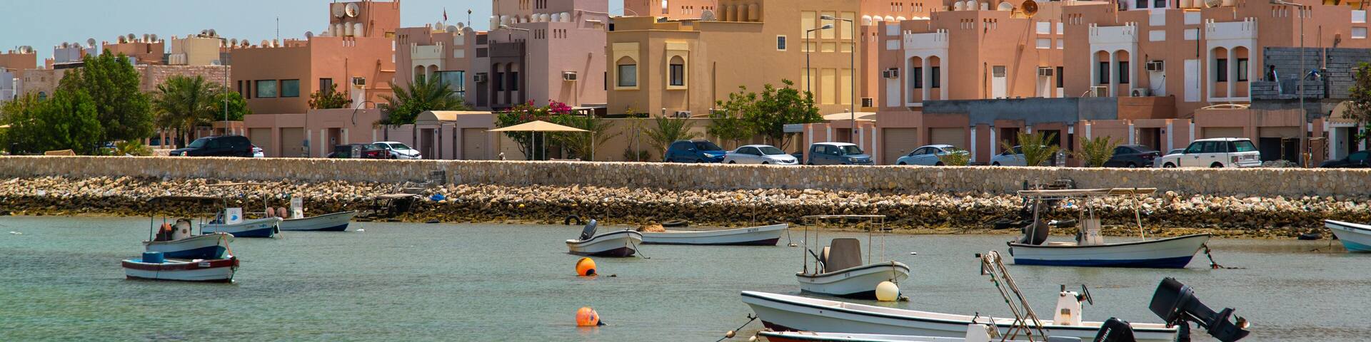 A coastal town with a prominent mosque, boats docked along the shore, and a stone seawall.